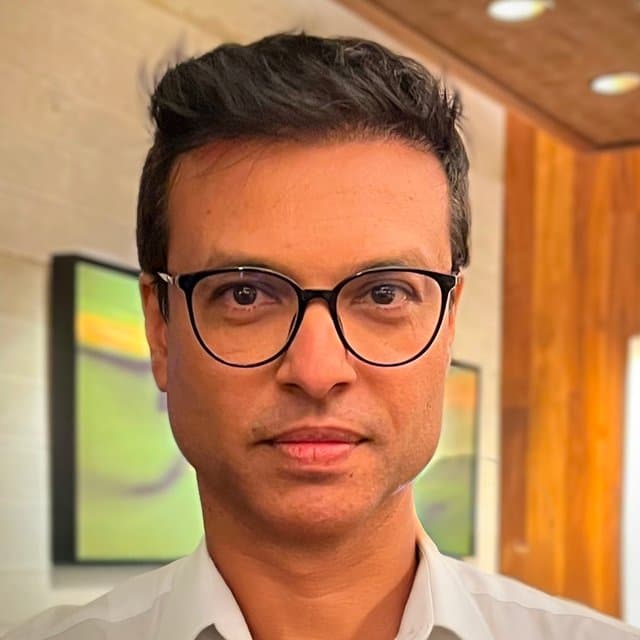 Prashant Palsokar, mid-shot portrait in a white shirt with dark-framed round glasses, photographed in warm interior light with abstract green-toned artwork behind him.