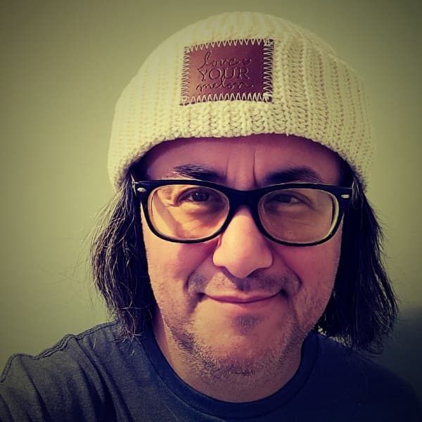 William Tincup, smiling slightly, wearing a cream beanie with a leather patch reading 'Love Your Melon' and dark-framed glasses, photographed in warm indoor light.