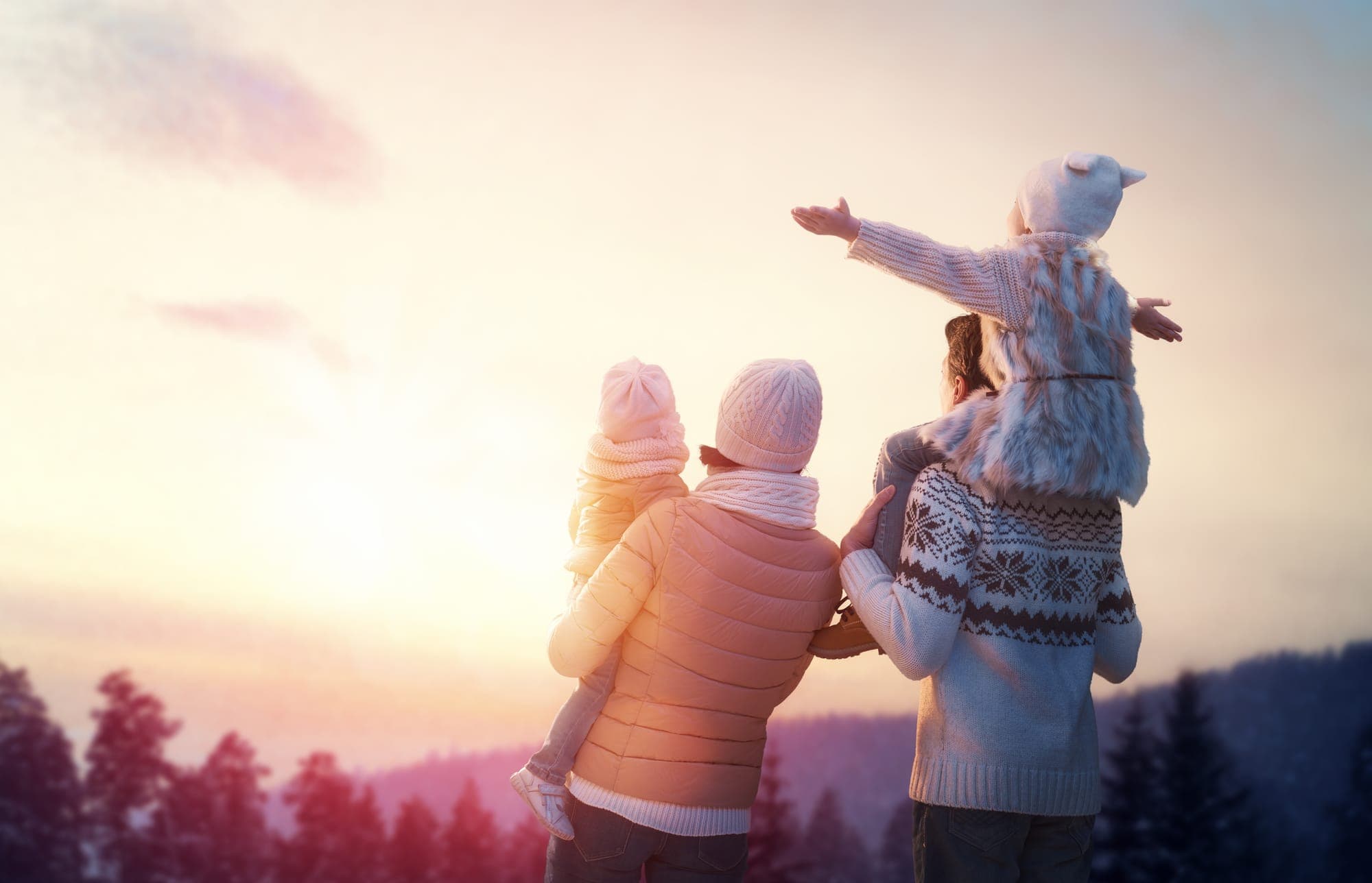 A family outdoors at sunset, parents carrying two young children, looking out at the horizon.